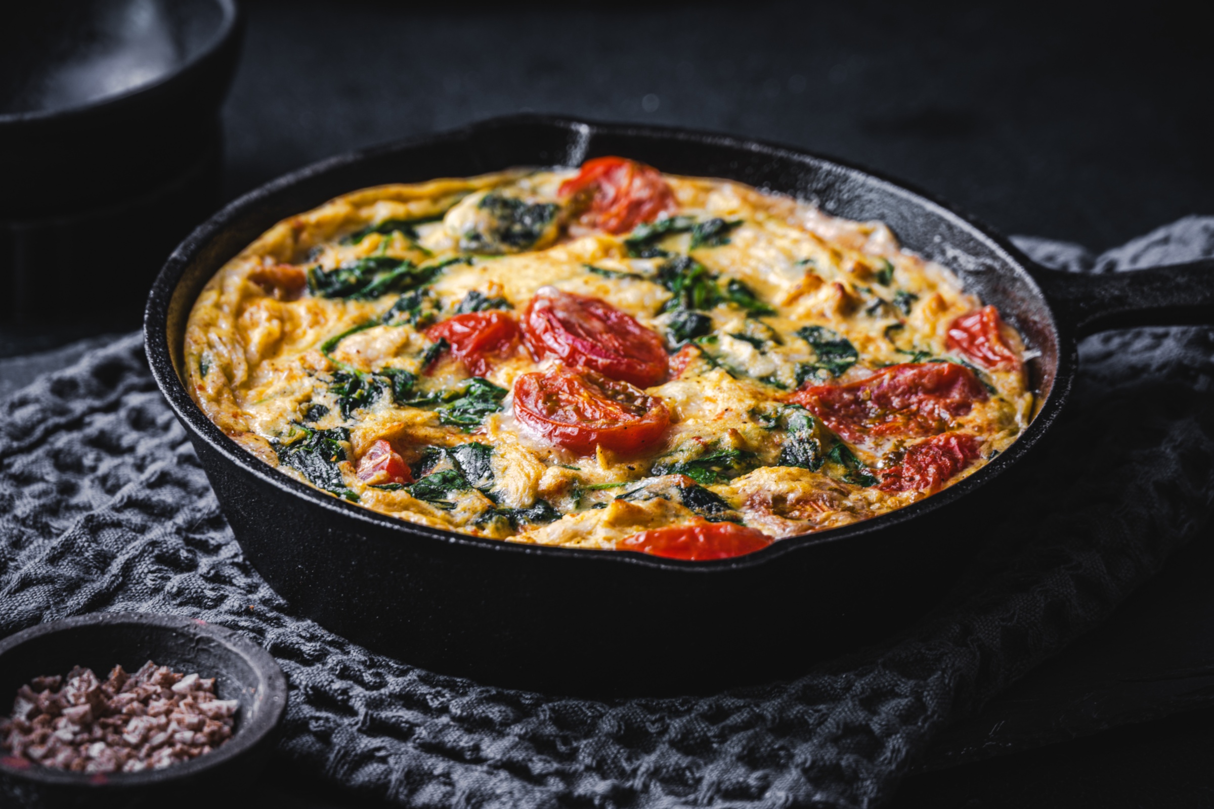 Überbackenes Gericht mit Spinat und Tomaten in einer gusseisernen Pfanne vor schwarzem Hintergrund.