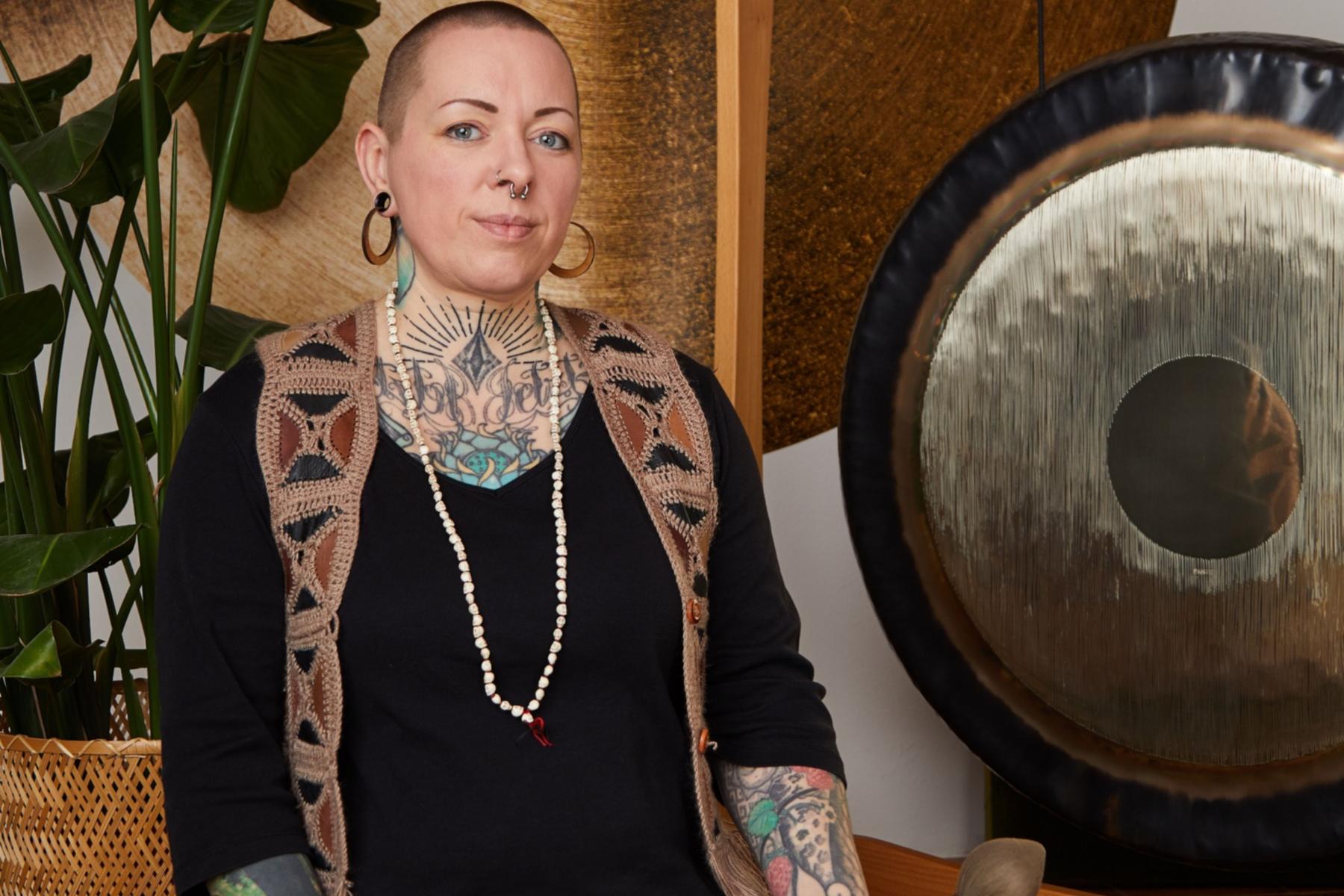 Die Dozentin sitzt mit gekreuzten Beinen auf dem Boden. Schräg hinter ihr steht ein großer Gong. Sie hält zwei Schlägel hierfür in der Hand.