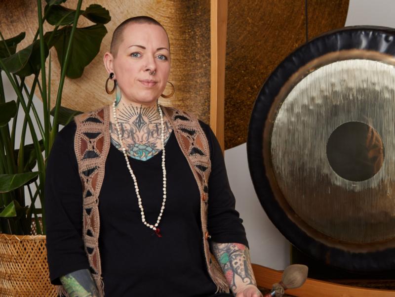 Die Dozentin sitzt mit gekreuzten Beinen auf dem Boden. Schräg hinter ihr steht ein großer Gong. Sie hält zwei Schlägel hierfür in der Hand.