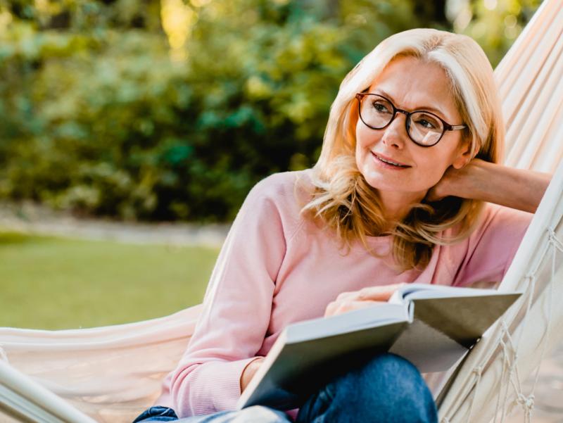 Eine Frau sitzt in einer Hängematte, hat ein aufgeschlagenes Buch auf ihren Beinen liegen und schaut in die Ferne.