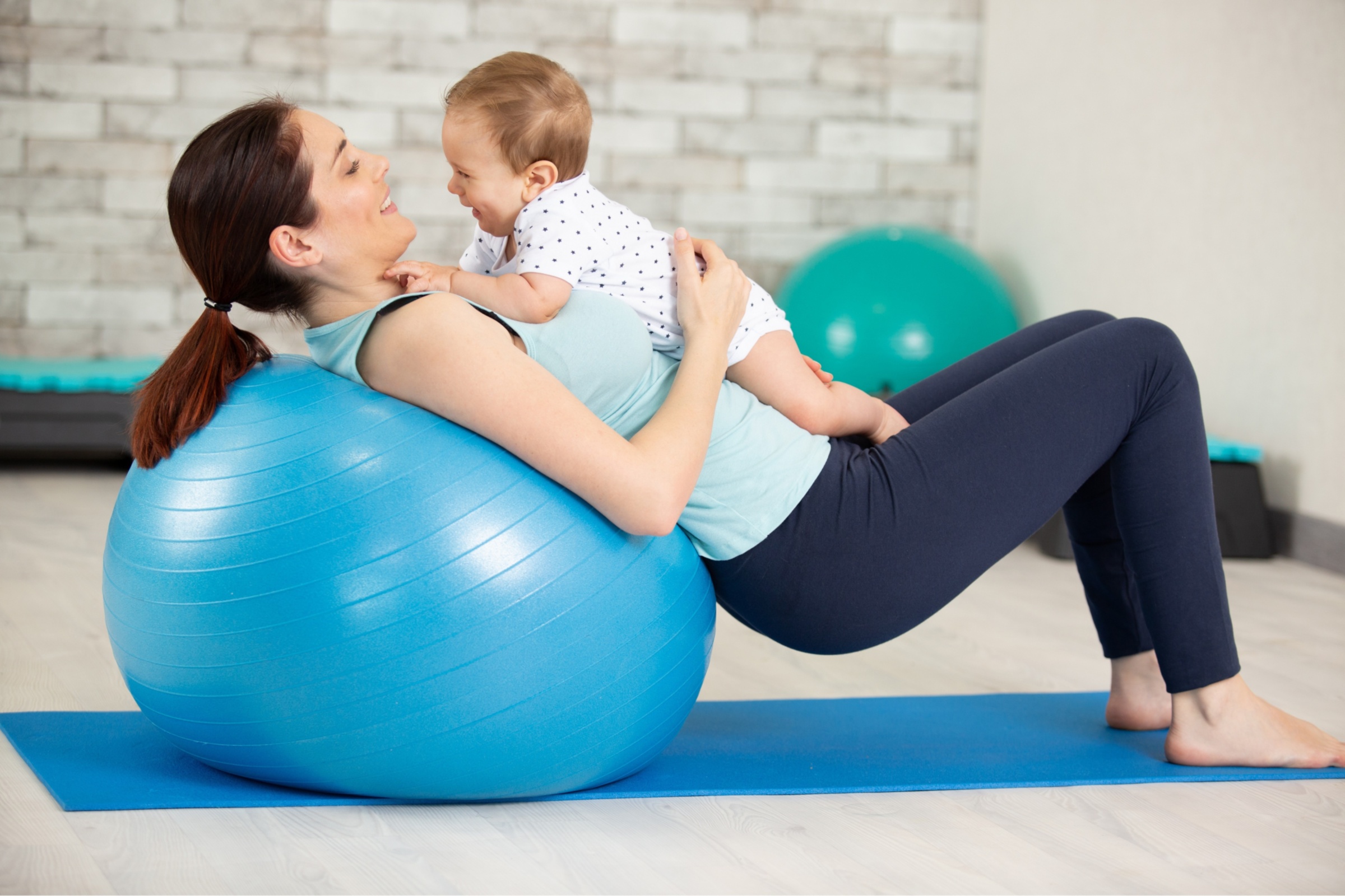Die Mutter liegt für eine sportliche Übung mit dem Rücken auf einem Gymnastikball. Ihr Baby liegt auf ihr. Beide schauen sich fröhlich an.