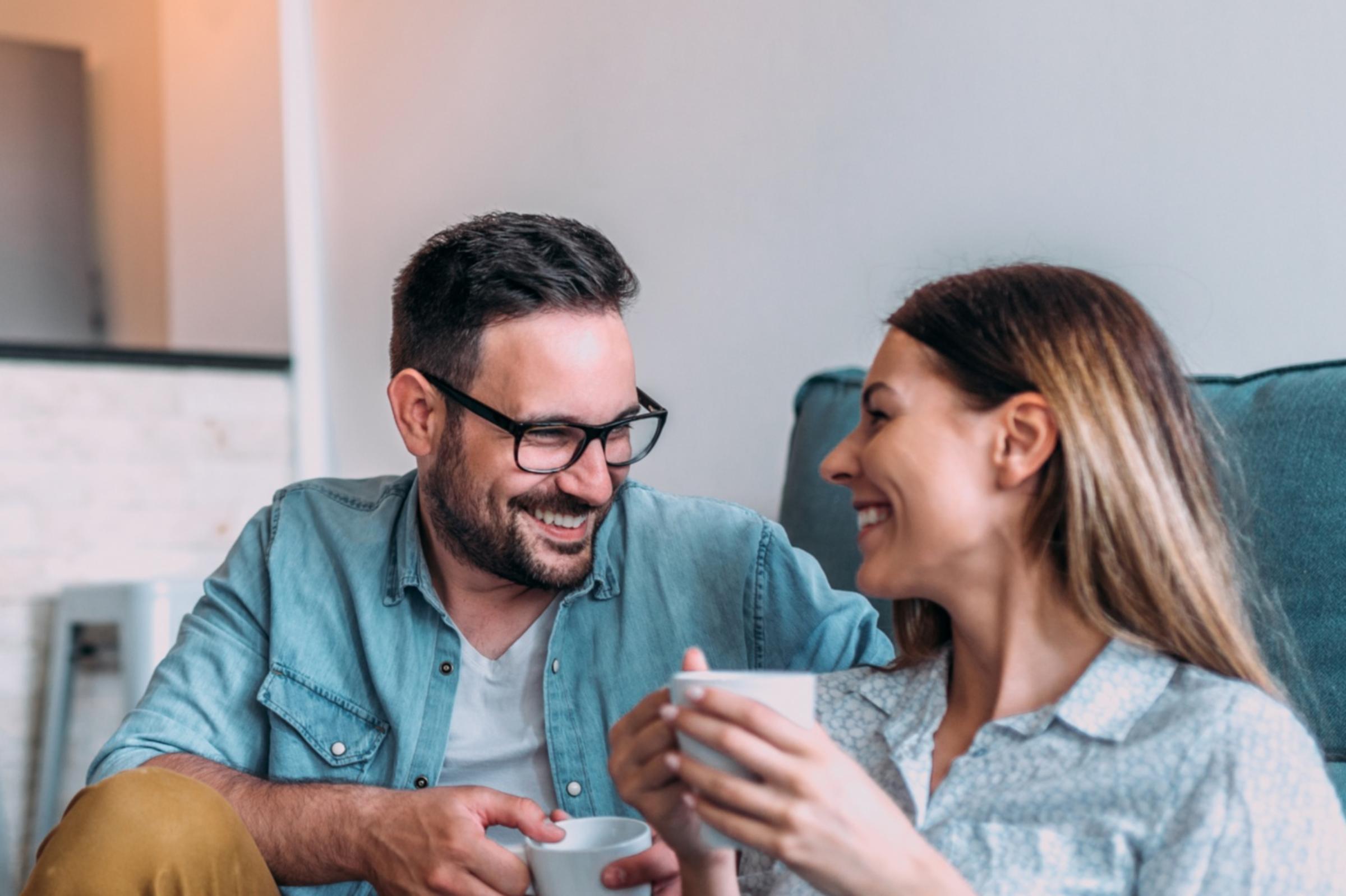 Eine Frau und ein Mann sitzen einander zugewandt auf dem Boden vor einem Sofa. Sie halten beide eine Tasse in der Hand.