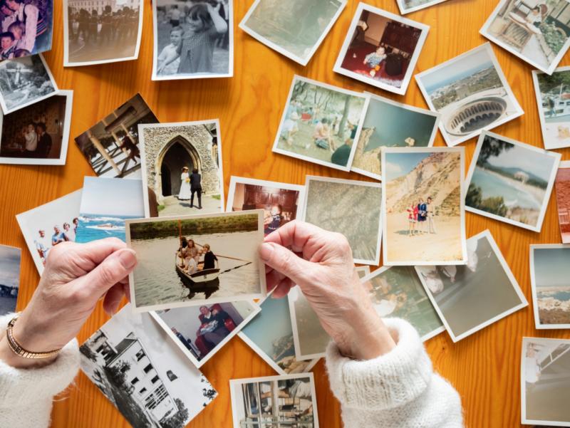 Bildausschnitt mit den Händen einer älteren Frau, die alte Familienfotos, ausgebreitet auf einem Holztisch, anschaut.