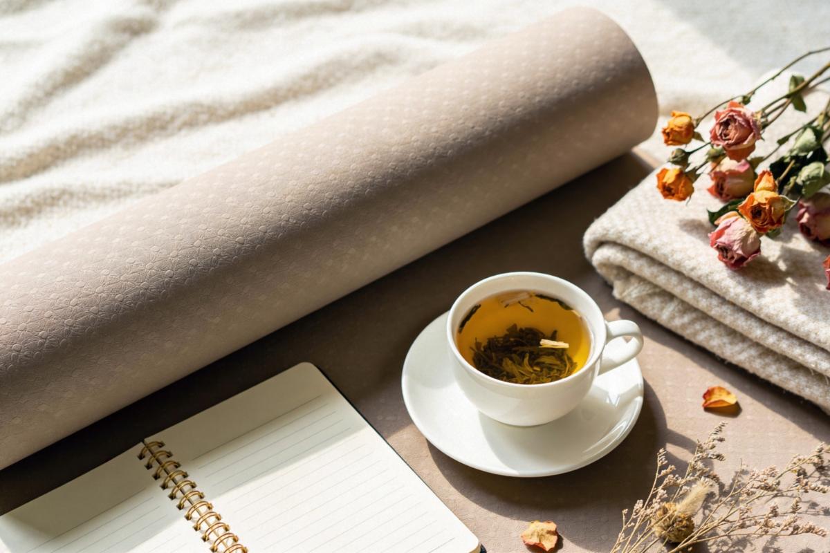 Auf einer Yogamatte stehen und liegen eine Tasse Tee, ein Schreibblock, getrocknete Blumen und ein Handtuch.