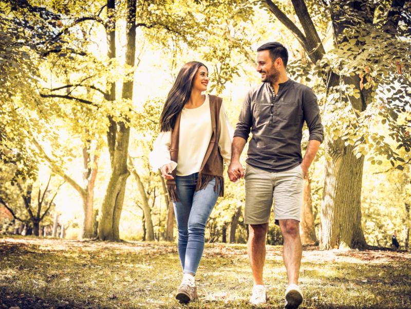 Eine Frau und ein Mann gehen einander zugewandt und an den Händen haltend durch einen herbstlichen Park spazieren.