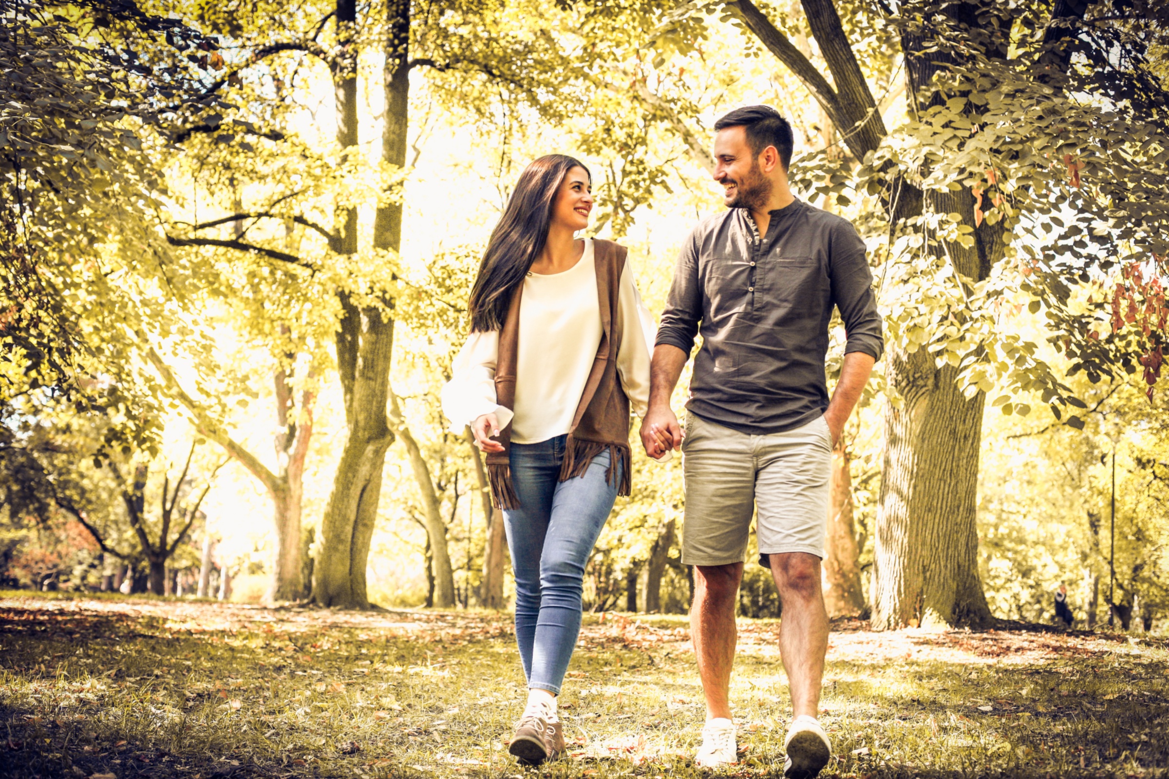 Eine Frau und ein Mann gehen einander zugewandt und an den Händen haltend durch einen herbstlichen Park spazieren.
